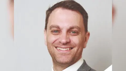 South Kesteven District Council A head and shoulders shot of a man smiling into the camera. He has short brown hair and is wearing a grey blazer and white shirt.