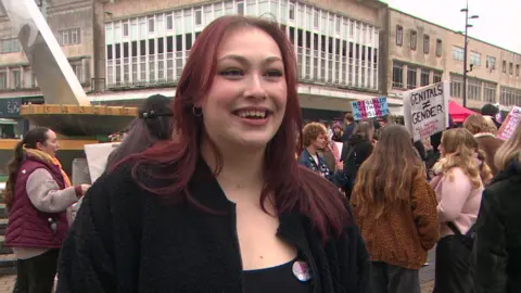 Close up of march lead Giuliette Alexandria. The organiser has mid-length red hair and is wearing a black top. She is standing in front of protestors who have gathered next to The Sundial in the city centre. 