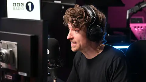 Greg James speaking into a black radio microphone with large black headphones over his head. He is talking and staring ahead which is to the left of the camera. There is a computer monitor in front of him to the right and a large BBC Radio 1 white sign to the left. There is a pink light behind him illuminating a sound board, which is slightly out of focus.