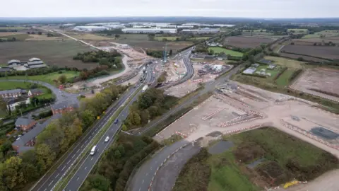 An aerial view of a construction site next to a dual carriageway surrounded by fields.