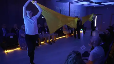 Students hold up a large yellow banner whilst an audience looks on in the new performing arts centre at Prior's Court.