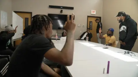 Rob White, wearing a black Hodia and a hat, stands next to the semester table where six other men sit. Many of them raise their hands.
