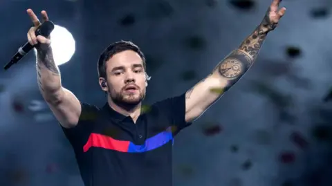 Getty Images Liam Payne holding a microphone wearing a black T-shirt with red and blue stripes 