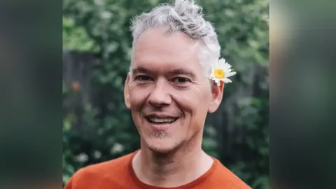 Tim Foskett Close up picture of Tim smiling looking at the camera. He has a white flower behind his ear. Tim has short grey hair and is wearing a orange t-shirt.
