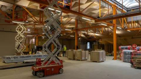 Construction work taking place in a former shop in Keighley. The building is empty with pillars supporting the roof alongside building materials and machinery with workers in the distance