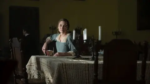 A woman playing Jane Austen sits at a table looking at the camera.
