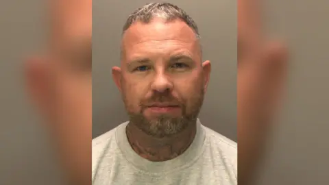 A police custody photograph of Michael Watling. He is wearing a grey jumper. Watling has short, greying hair and a short beard. His neck is also covered in tattoos.