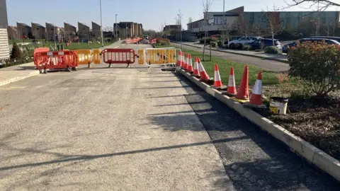 Shariqua Ahmed/BBC A road under construction. The grey, concrete road has orange and white cones on either side. There are orange and yellow barriers in the distance.