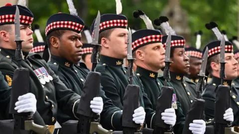 Eight soldiers from the Royal Regiment of Scotland standing in a line in dark ceremonial uniforms. They are all wearing white gloves and holding rifles. 