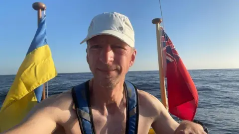 Leo Krivskiy Man on boat with sea surrounding he has no top on a blue harness is over his shoulders - and is wearing a white baseball cap.  He is looking at the camera.