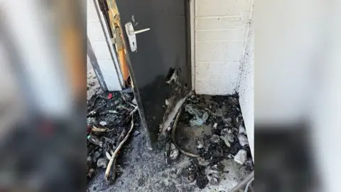 Burnt items are lying on a floor next to a door. They are black and charred. The white wall has black specks on it.