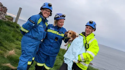 Eyemouth Coastguard Rescue Team Two people in blue outdoor wear beside a man in a high vis jacket who is holding a dog wrapped in a blanket