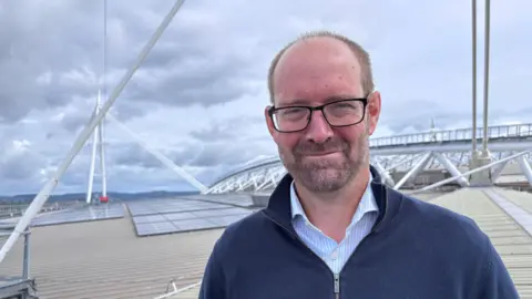 Gavin Marshall stands on the roof of the stadium. He has short, light brown hair, is wearing black glasses and a navy blue sweater