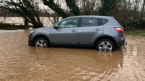 A dark silver car with murky floodwater halfway up its wheels. There is floodwater all around and some trees and bushed in the background.