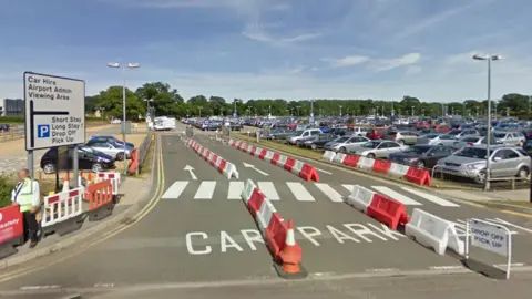 Google A car park filled with cars. White painted signs on the road read "car park". There are temporary white and orange plastic barriers at the entrance. There is also a sign which reads "Drop off, pick up".