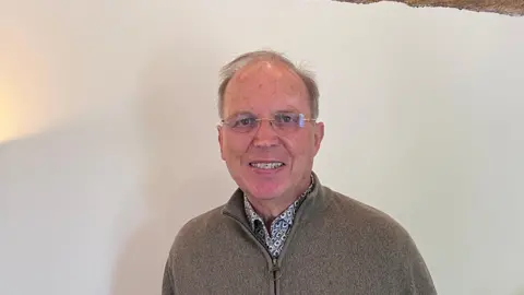 Dr Roger Cliffe pictured in a front of a white wall. He has short brown hair and is wearing thin glasses. He is also wearing a patterned shirt and a beige fleece.