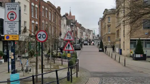 Google Gloucester city centre. The view facing up the high street. There are various signs for drivers including a speed sign and pedestrian warning. There are black bollards and a black railing to allow pedestrians to walk safely on either side of the street next to shops.