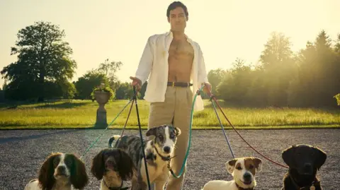 Disney+ actor Alex Hassell as Rupert Campbell-Black in the Disney+ series Rivals, walking dogs with his shirt undone to show his chest. 