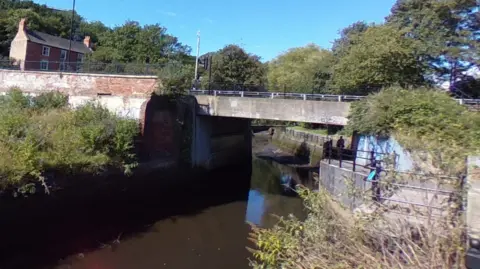 Google Photo of a low concrete bridge over small river on clear blue day. There is a large house in the background and the area is surrounded by trees and bushes. 