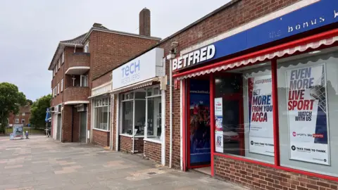 Shaun Whitmore/BBC A betting shop is shown to the right of the picture in the foreground, with an empty pavement to the left. A tech repair shop is alongside it and then a three-storey flat block looms in the background.