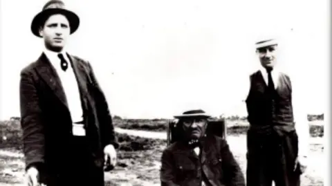 Josephine Smith A black and white picture of three men in a field wearing hats and suits. Two younger men are standing either side of an older man who is sitting down.