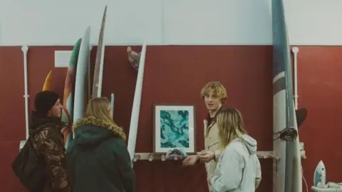 Ebbi Rose Jacob Arnold showing people surfboards he has made. His girlfriend India is stood next to him and one of her textile designs is visible behind them. Surfboards line the walls in multiple colours and shapes. 