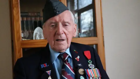 A man with short white hair wears a blue hat with a blue jacket. The jacket is covered with ribbons and badges and war medals. He wears it over a light blue shirt and a red and blue and white striped tie. Behind him is a mirror in a wooden frame.