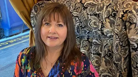 Supplied Councillor Kerry Robinson Payne, wearing a multicoloured blouse, sitting on a luxurious train. She is smiling, and has long brunette hair. The seat fabric is highly patterned.