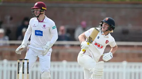 Jordan Cox appears to be in discomfort after playing a shot for Essex, while Somerset wicketkeeper James Rew stands behind the stumps