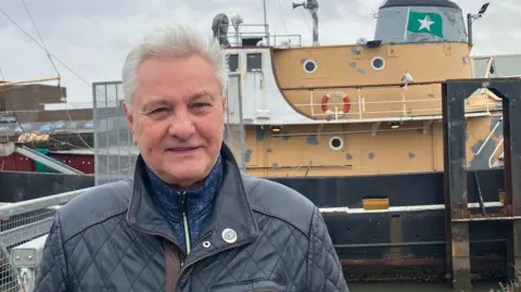 A man with short grey hair smiles as he stands in front of 1950s trawler at the side of a dock. He wears a black biker-style leather jacket and a blue tracksuit top. The trawler has a black hull, with white and tan-coloured upper works and a grey funnel, which has a green flag painted on it with white star emblem.