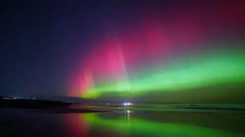 Colin Conti/BBC Weather Watchers The aurora appears as pink, green and white and the display is reflected in the sea.