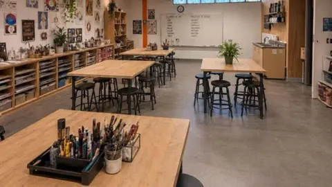 LDRS A modern-looking classroom with four wooden tables and black stools, with pens and paintbrushes on the tables and art supplies around the edge of the room. 