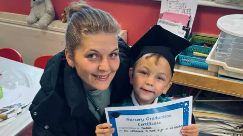 Family photograph A woman with her hair tied back smiles as she leans next to a small boy in a black cap holding a 'nursery graduation certificate'. Both are smiling and appear to be in a school. 