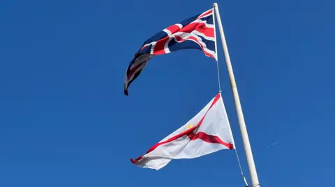 A picture of two flags flying high from a flagpole. There is one flag above the other.