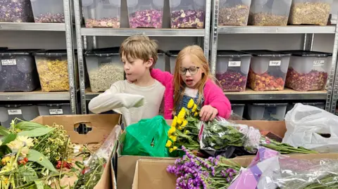 A girl and boy lean over boxes of bunches of flowers. The girl on the right has long red hair, glasses, a pink fluffy cardigan, a pink t-shirt and denim dungarees. She is talking whilst touching a bunch of yellow flowers. Her other arm is around the boy. The boy has short fair hair, a long sleeved white top and is looking at a bunch of flowers. Behind them are rows of shelves with labelled transparent boxes full of different coloured flower petals.