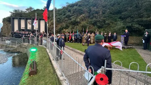 People gathered at the Heroes on the Water base at Port Soderick for the ceremony. Veterans and representatives from the armed forces and emergency services stand with their ceremonial flags and standards. Lieutenant Governor Sir John Lorimer is standing on a platform next to the bench, talking into a microphone.