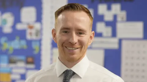 Jamie Niblock/BBC Head of school Julien Mealey in front of a blue board in a classroom which has work displayed on it. He has short ginger hair and stubble and is smiling while wearing a white shirt and dark tie.