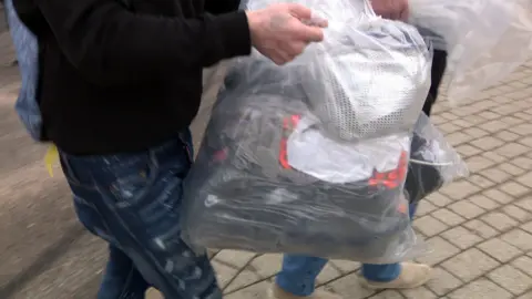 An unidentified man wearing jeans and a black jumper carries a plastic bag of belongings.