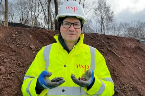 A man wearing a hi-vis jacket and white hard hat with a logo saying WSP on both. He is holding two pieces of shale, one burnt and one not.