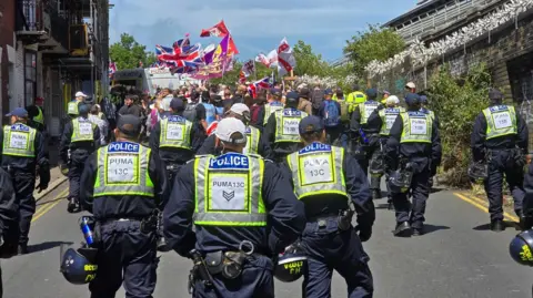 Sussex Police A group of police officers standing in front of a group of protesters who are holding a variety of flags, including the flag of St George and Union Jack. 