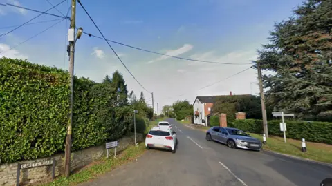 Google A rural crossroads showing a tall hedge above a stone wall to the left, and a street sign saying CAREYS ROAD. There are cars parked on the grass verge next to the hedge. Another car is travelling towards the camera and is about to pass an old-style road sign. There is a white-rendered two storey house in the distance and a tree to the right.