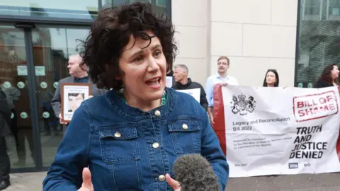 PA A woman with wiry black hair speaking into a mic, she has a denimn jacket on. Behind her is protestors with a white banner. 