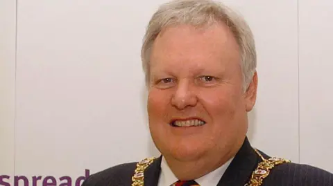 Pacemaker Lord Browne standing smiling at the camera. He is wearing a mayoral chain, a black suit and a tie with red, blue and yellow stripes. 