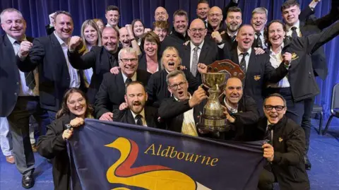 4barsrest A group of people dressed smartly in suits gathered for the camera all smiling or looking thrilled as two in front hold a large trophy and others hold an Aldbourne flag.