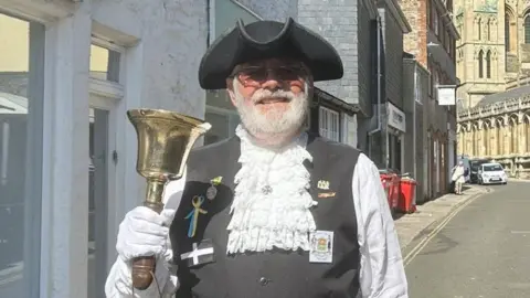 A town crier in ceremonial dress including a black hat and white laced shirt holds a gold bell and smiles at the camera in the street. 