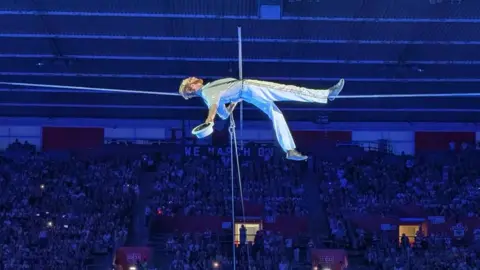 Graham Harries Ellis holds his white top hat in his right hand, and lies flat along a tight rope. The background shows large crowds of people watching on from the stadium seating. 
