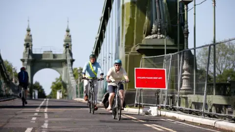Getty Images Cyclists riding across the deck of Hammersmith Bridge, passing a red sign on a metal barrier that reads "cyclists keep left" with the green suspension towers visible in the background.