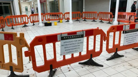 The inside of a shopping centre - the image shows some orange barriers which have fenced off a section of the flooring, for maintenance work. All the shops are shut and two people are wandering around in the background.