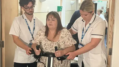 Image of Sonia walking with an aid being supported by two hospital physios in white uniforms.