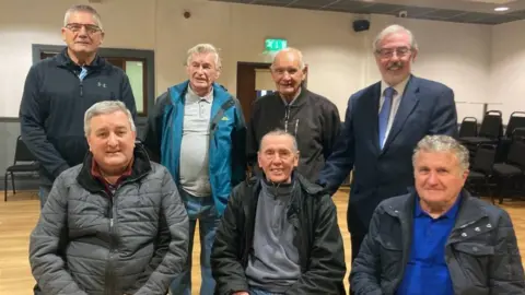 A group of seven men standing for a group photo in a hall